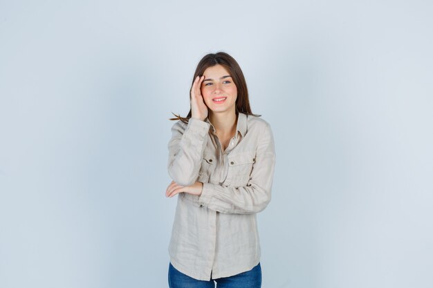 Jovencita manteniendo la mano en la cabeza mientras sonríe en casual, jeans y luce alegre. vista frontal.