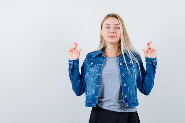 Jovencita mostrando los dedos cruzados en camiseta, chaqueta vaquera, falda y luciendo linda