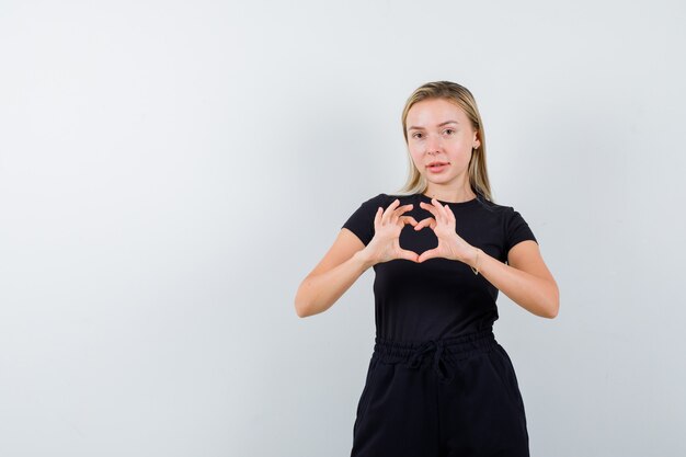 Jovencita mostrando gesto de corazón en camiseta, pantalón y luciendo lindo, vista frontal.