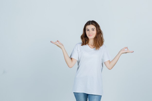 Jovencita mostrando gesto de meditación en camiseta, jeans y mirando pacífica, vista frontal.