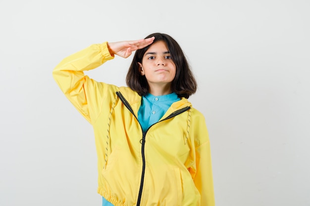 Jovencita mostrando gesto de saludo en chaqueta amarilla y mirando confiado, vista frontal.