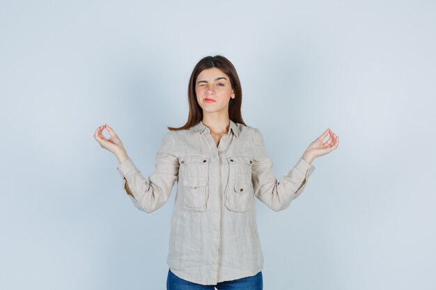 Jovencita mostrando gestos de yoga mientras parpadea en casual, jeans y parece pacífica. vista frontal.