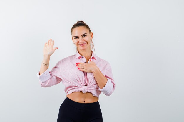 Jovencita mostrando la palma, manteniendo la mano en el pecho en camisa, pantalones y mirando alegre, vista frontal.