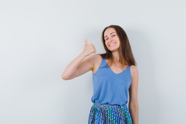 Jovencita mostrando el pulgar hacia arriba en camiseta, falda y mirando feliz, vista frontal.