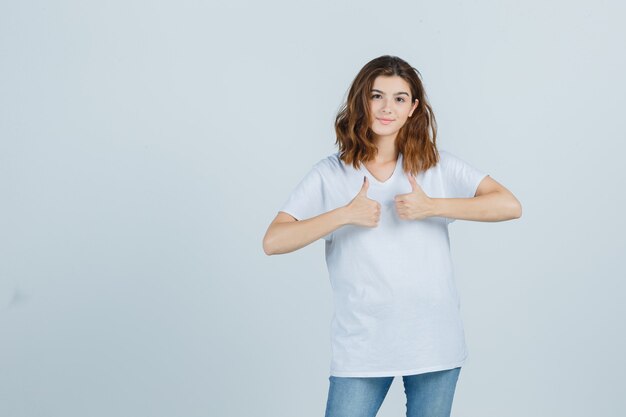 Jovencita mostrando los pulgares para arriba en camiseta, jeans y mirando alegre, vista frontal.