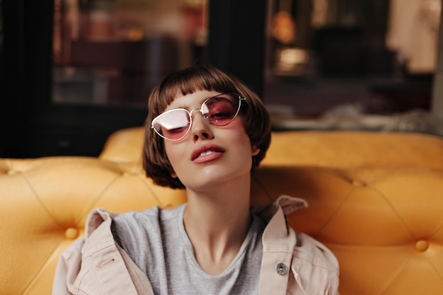 Jovencita con el pelo corto posando en el sofá dentro Encantadora mujer morena con gafas de sol rosadas y traje de denim beige mirando a la cámara en el café