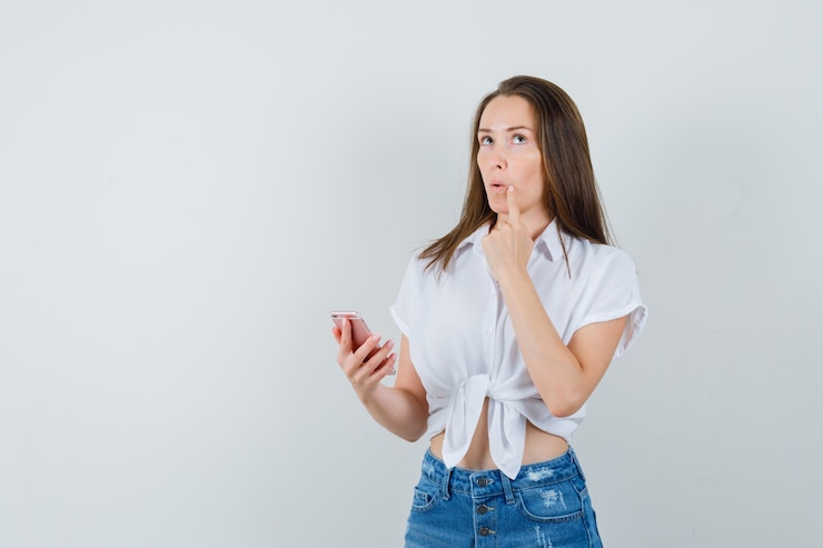 Foto gratuito jovencita pensando en algo mientras sostiene el dispositivo en una blusa blanca y mira pensativa, vista frontal. espacio para texto