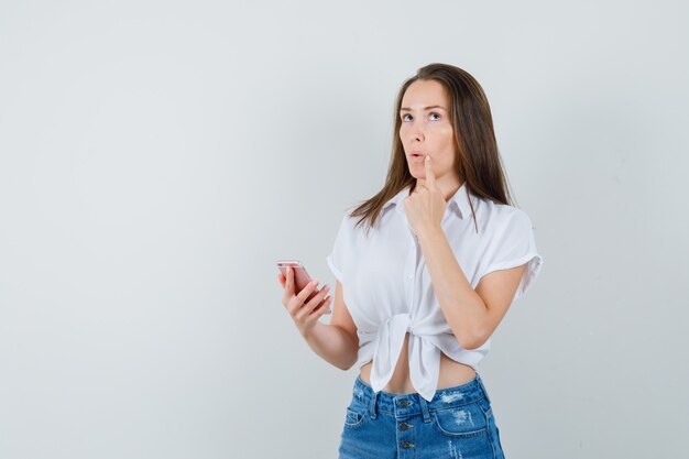 Jovencita pensando en algo mientras sostiene el dispositivo en una blusa blanca y mira pensativa, vista frontal. espacio para texto