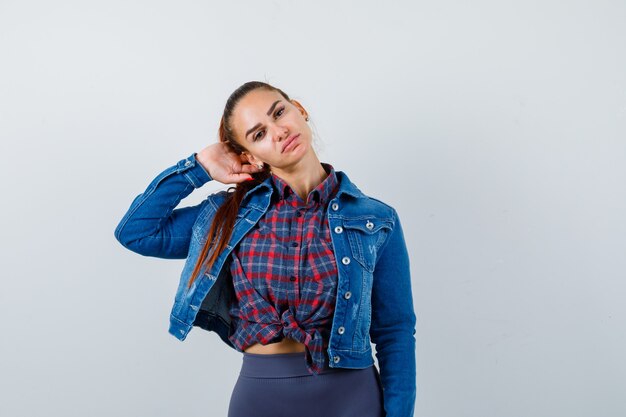 Jovencita posando con la mano detrás de la cabeza en camisa a cuadros, chaqueta de mezclilla y mirando confiado, vista frontal.