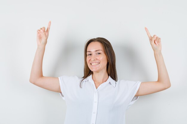 Jovencita posando mientras apunta hacia arriba con una blusa blanca y se ve alegre.