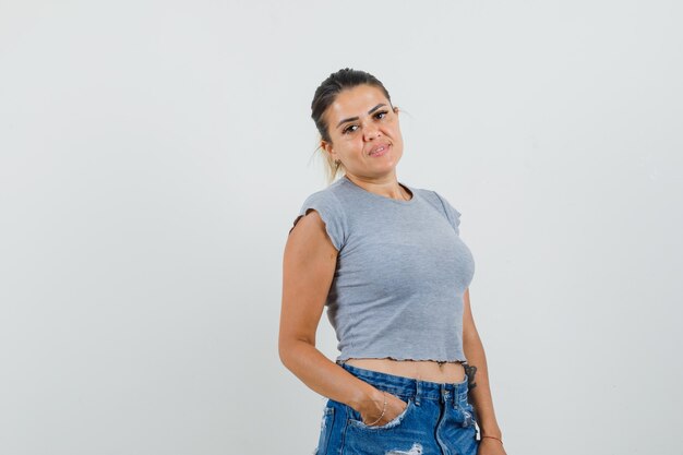 Foto gratuita jovencita posando mientras sostiene la mano en el bolsillo en una camiseta, pantalones cortos y parece cautivadora