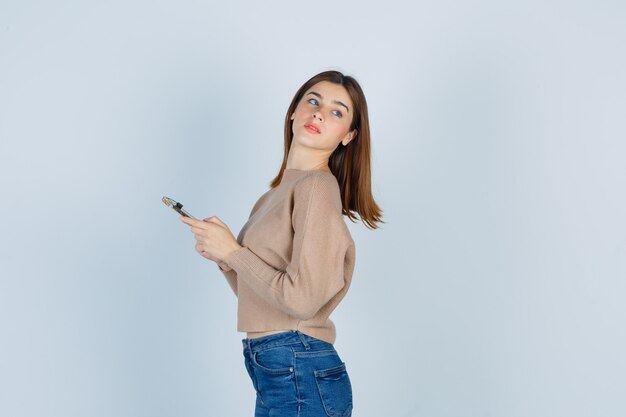 Jovencita posando mientras sostiene el teléfono móvil en suéter beige, jeans y luce atractivo. vista frontal.