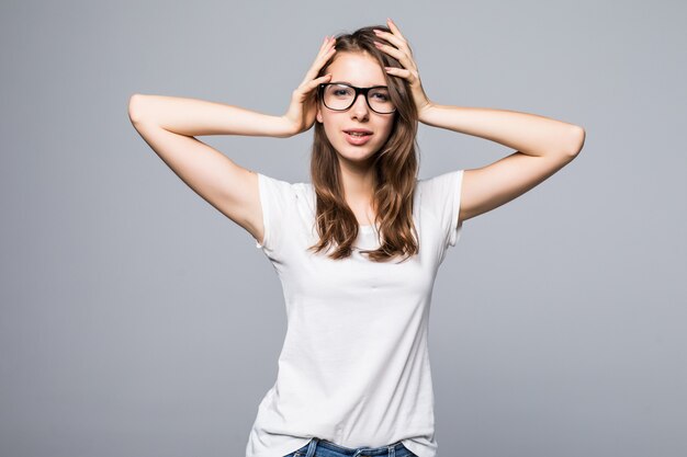 Jovencita sexy con gafas en camiseta blanca y jeans azul sostiene sus brazos en el cabello frente a fondo blanco de estudio