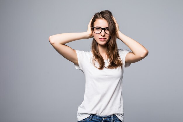 Jovencita sexy con gafas en camiseta blanca y jeans azul sostiene sus brazos en el cabello frente a fondo blanco de estudio