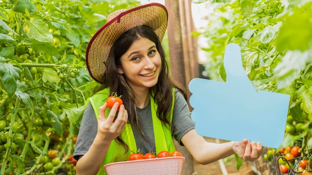 Jovencita sonriente sosteniendo un cartel azul y tomate maduro en el invernadero