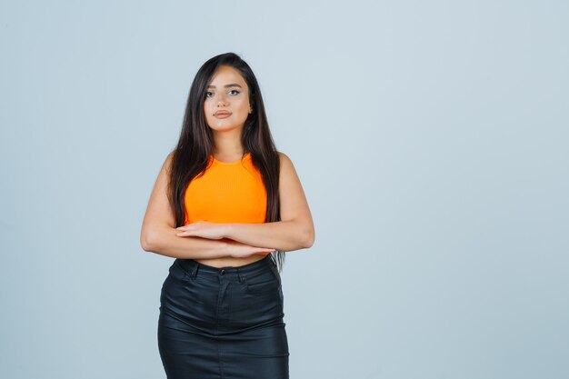 Jovencita sosteniendo los brazos juntos cerca del estómago en camiseta, minifalda y mirando confiada, vista frontal.