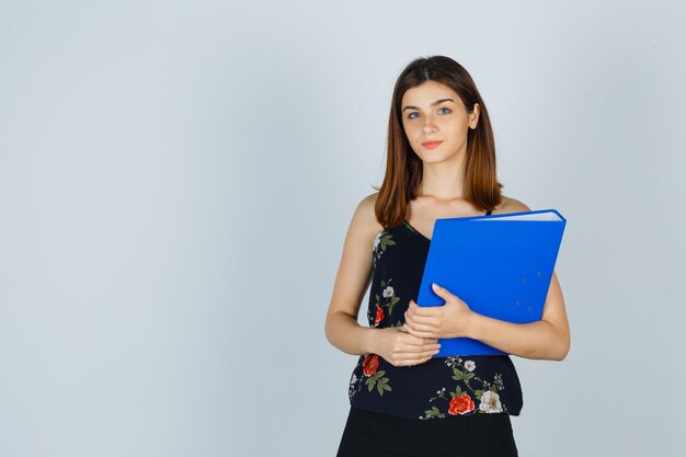 Jovencita sosteniendo una carpeta en blusa, falda y luciendo optimista. vista frontal.