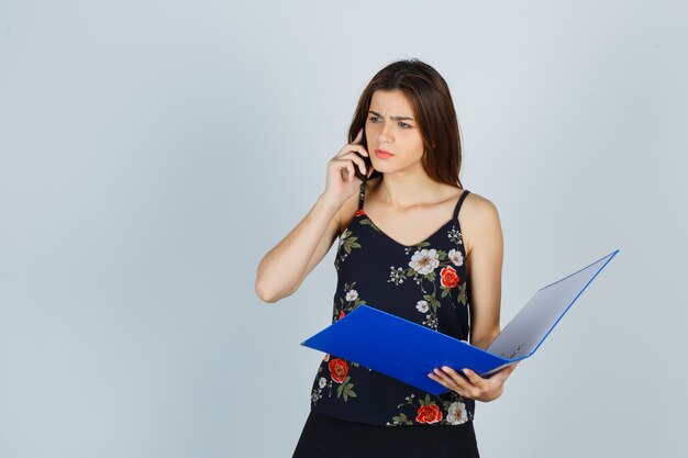 Jovencita sosteniendo una carpeta, hablando por teléfono móvil en blusa y mirando confundido. vista frontal.