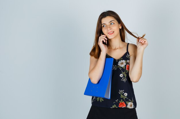 Jovencita sosteniendo una carpeta, hablando por teléfono móvil, girando el cabello alrededor de sus dedos en blusa, falda y mirando alegre. vista frontal.