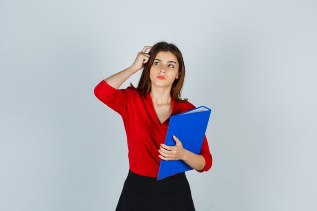 Jovencita sosteniendo la carpeta mientras se rasca la cabeza en blusa roja, falda y mirando pensativo