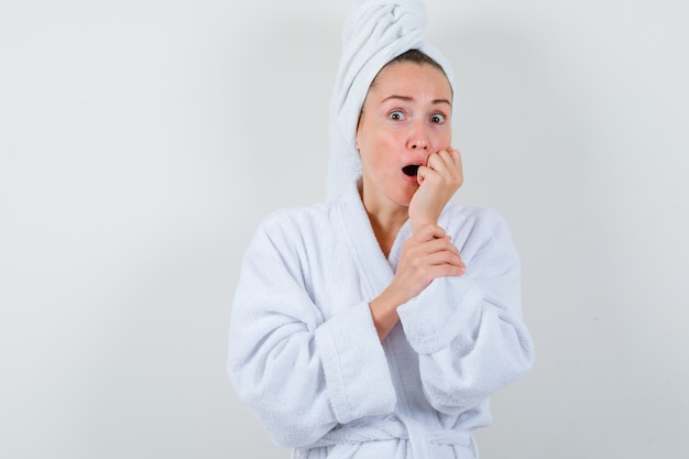 Jovencita sosteniendo el puño cerca de la boca en bata de baño blanca, toalla y mirando sorprendido, vista frontal.