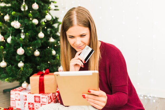 Jovencita con tableta y tarjeta de plástico cerca de cajas de regalo y árbol de navidad