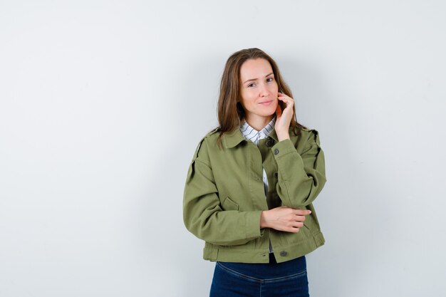 Jovencita tocando la piel de la cara en la mejilla en blusa, chaqueta y luciendo elegante, vista frontal.