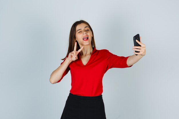 Jovencita tomando selfie mientras muestra el signo de la victoria en blusa roja