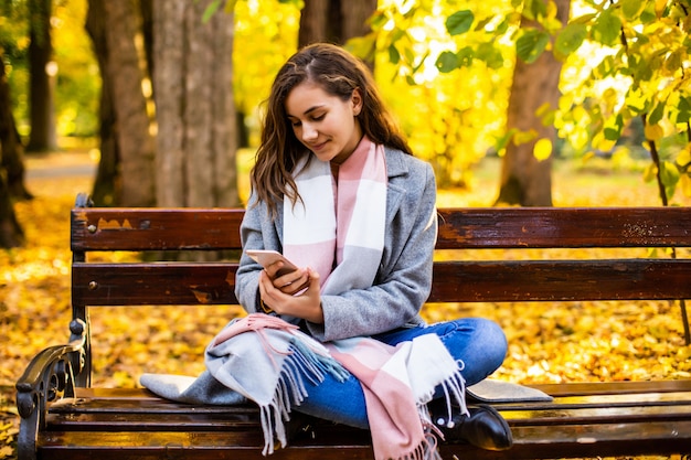 Jovencita usando un teléfono inteligente y mensajes de texto sentado en un banco de un parque urbano de otoño