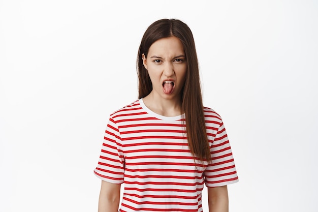 Las jóvenes adolescentes muestran aversión, extienden la lengua, fruncen el ceño y entrecierran los ojos por disgusto, aversión, mal gusto de algo horrible, de pie en camiseta contra fondo blanco.