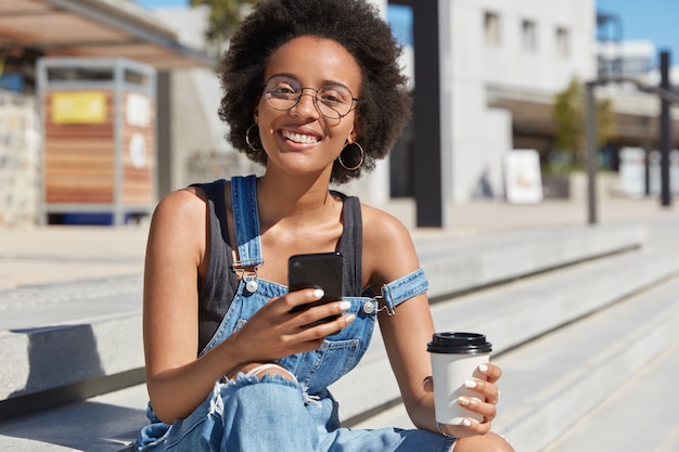 Jóvenes adolescentes de piel oscura positivos mensajes en línea, disfrutan del café para llevar de una taza desechable, usan overoles de mezclilla andrajosos, disfrutan del tiempo libre al aire libre. Estilo callejero. Concepto de estilo de vida.