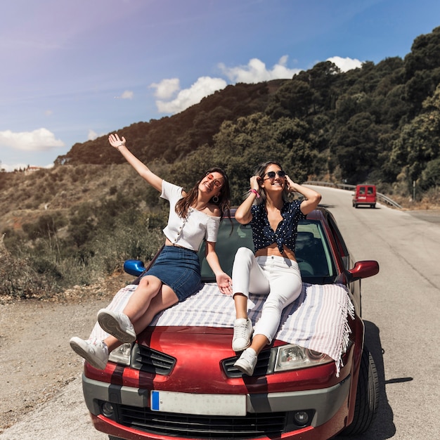 Jóvenes amigas sentada sobre el capó del coche burlarse de carretera