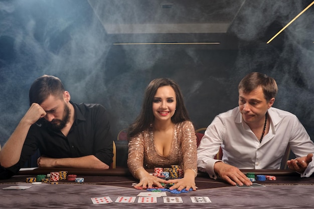 Foto gratuita jóvenes amigos alegres están jugando al póquer en el casino en humo. la chica ha ganado y los chicos han perdido. los jóvenes están haciendo apuestas esperando una gran victoria. juegos de azar por dinero. juegos de fortuna.