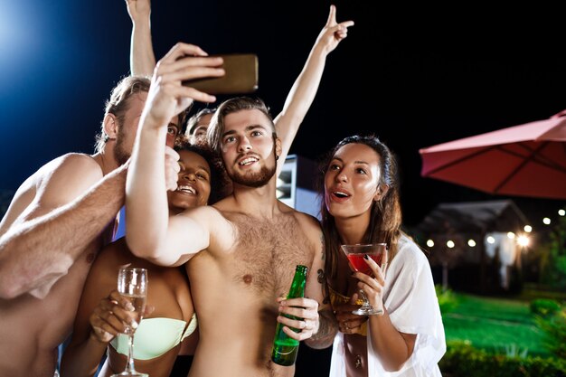 Jóvenes amigos alegres sonriendo, regocijándose, haciendo selfie, descansando en la fiesta