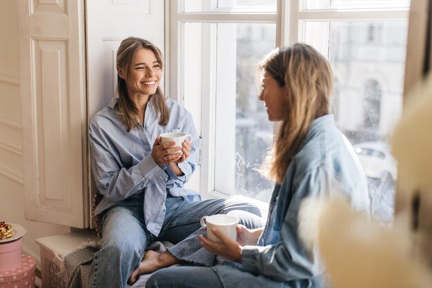 Jóvenes amigos caucásicos se reúnen en la ventana con vista a la ciudad, hablan, charlan y beben té. Concepto de fin de semana feliz