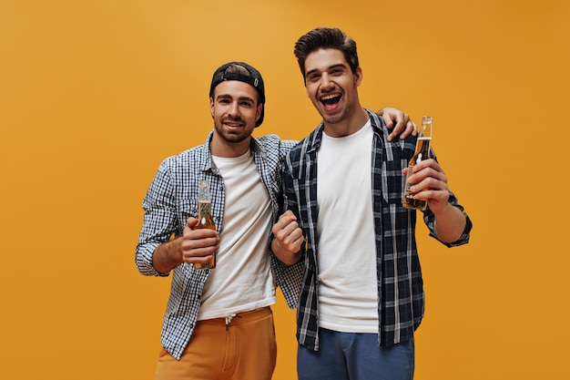 Jóvenes amigos geniales felices sostienen botellas de cerveza sobre fondo naranja Los hombres encantadores en camisas a cuadros se regocijan en aislados