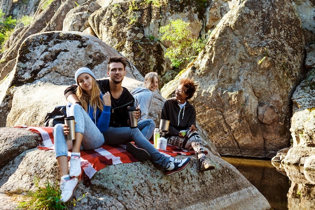 Jóvenes amigos sentados en roca en el cañón, sonriendo, bebiendo té