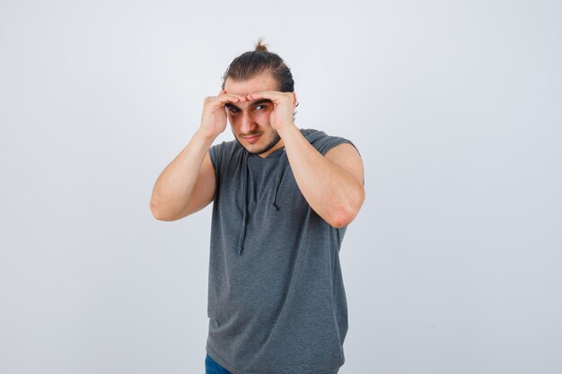 Los jóvenes aptos para hombres tomados de la mano en la cabeza para ver claramente en una sudadera con capucha sin mangas y mirando enfocado, vista frontal.