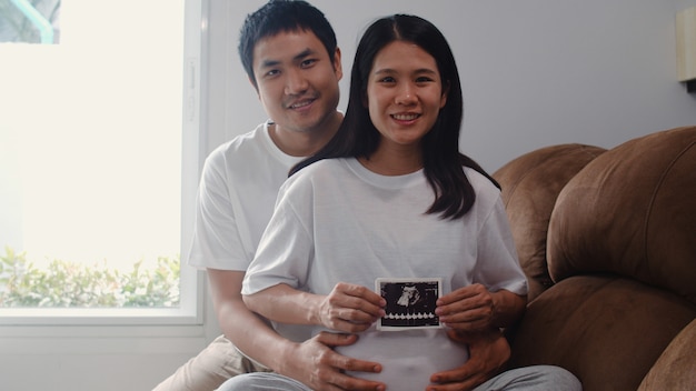 Jóvenes asiáticos embarazadas pareja muestran y mirando la foto de ultrasonido bebé en el vientre. Mamá y papá se sienten felices sonriendo pacíficos mientras cuidan al niño acostado en el sofá en la sala de estar en casa.