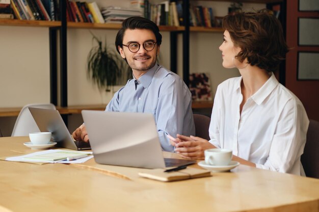 Jóvenes y atractivos colegas de negocios se miran felizmente mientras trabajan juntos en una laptop en una oficina moderna