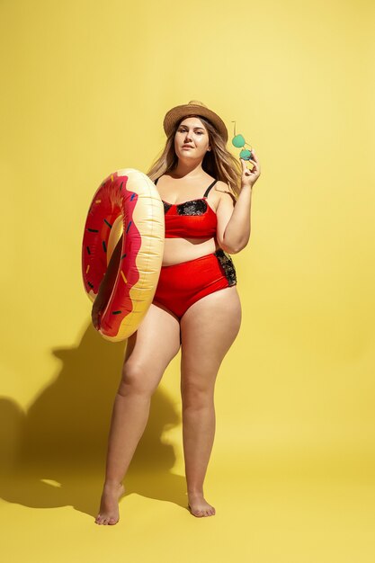 Jóvenes caucásicos de talla grande modelo femenino preparando vacaciones en pared amarilla. Mujer en traje de baño rojo, sombrero y gafas de sol.