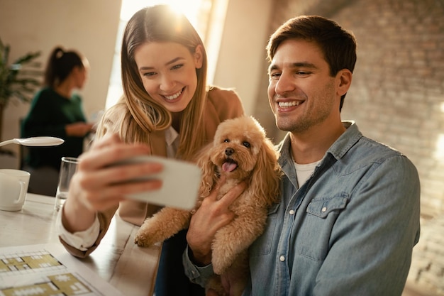 Foto gratuita jóvenes colegas felices divirtiéndose mientras toman selfie con un caniche en la oficina