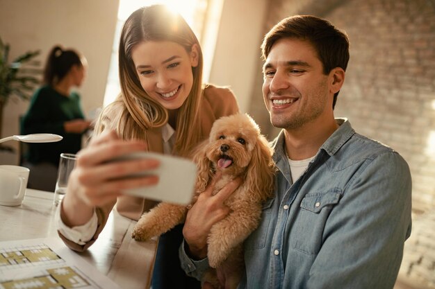 Jóvenes colegas felices divirtiéndose mientras toman selfie con un caniche en la oficina