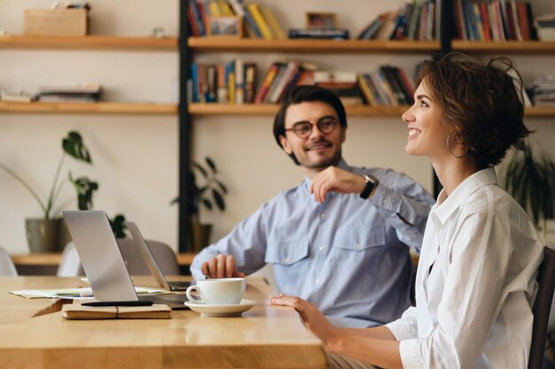 Jóvenes colegas de negocios atractivos sentados en la mesa con laptop y café mientras hablan felizmente en el trabajo en una oficina moderna