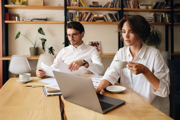 Jóvenes colegas de negocios atractivos trabajando cuidadosamente en un nuevo proyecto sentados en la mesa con una computadora portátil y papeles en una oficina moderna