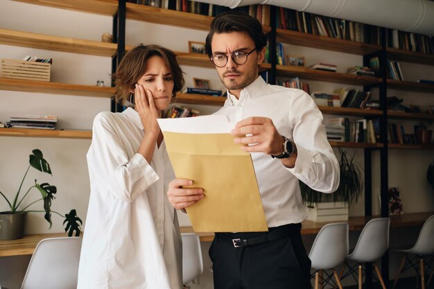 Jóvenes colegas de negocios leyendo cuidadosamente la carta del sobre mientras trabajan juntos en la oficina moderna