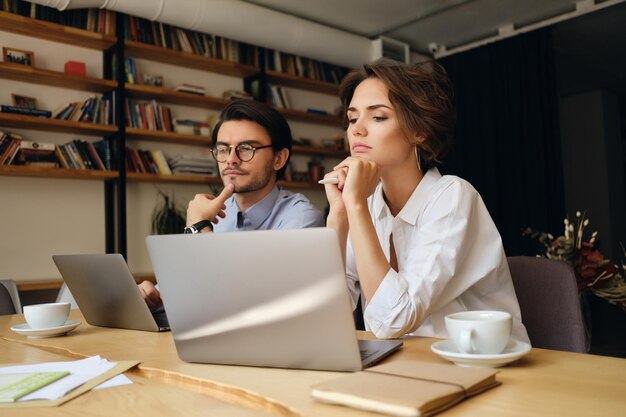 Jóvenes colegas de negocios pensativos sentados en el escritorio mientras trabajan cuidadosamente en una computadora portátil con café en una oficina moderna