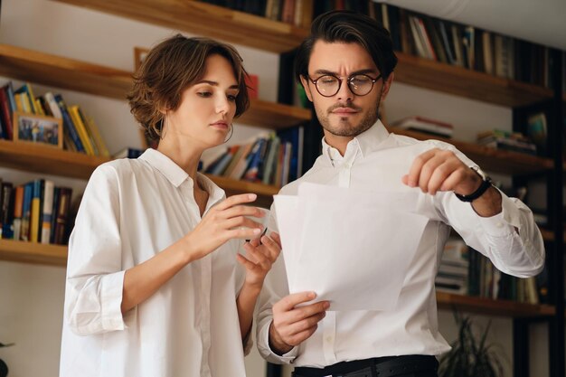 Jóvenes colegas de negocios reflexivos de pie con papeles y discutiendo un nuevo proyecto juntos en una oficina moderna