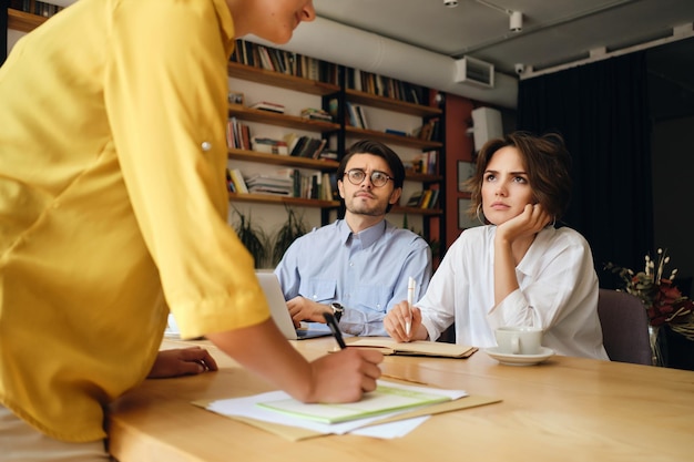 Jóvenes colegas de negocios sentados en el escritorio con una computadora portátil mirando cuidadosamente al jefe mientras discuten el trabajo juntos en la oficina moderna