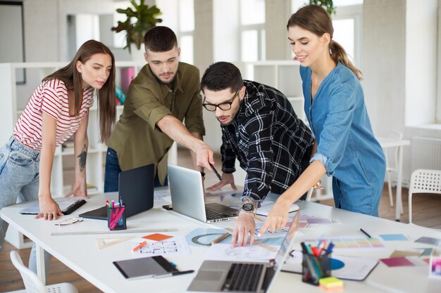 Jóvenes creativos con laptop y bocetos discutiendo juntos un nuevo proyecto Grupo de chicos y chicas trabajando en una oficina moderna y acogedora